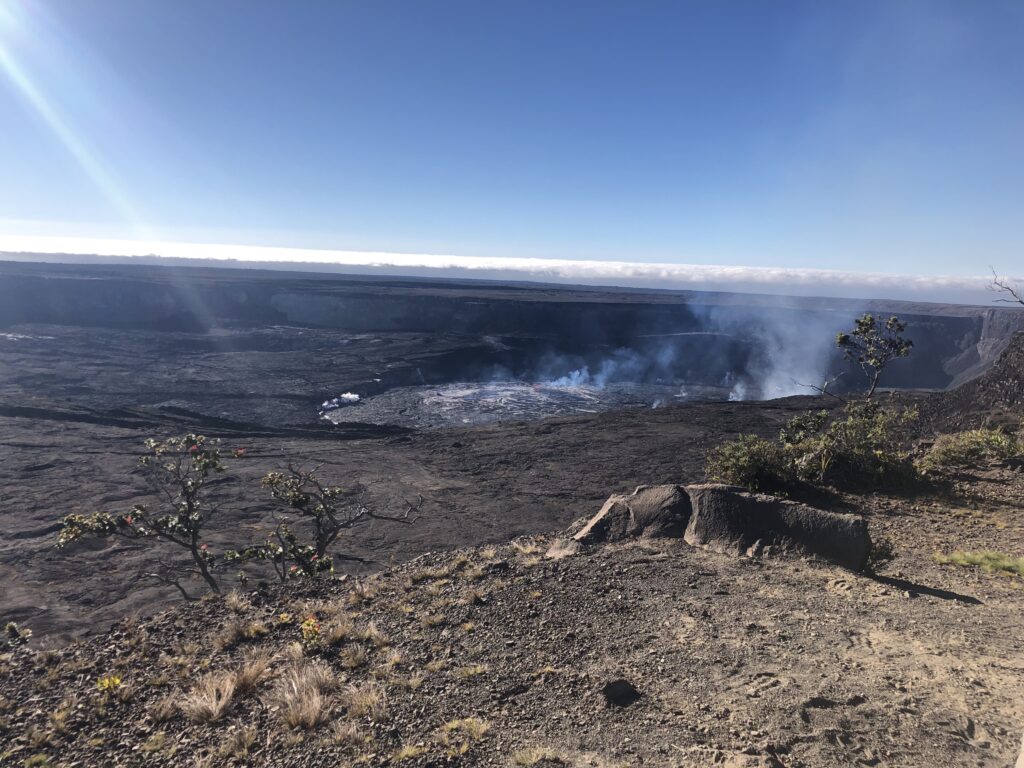 Kīlauea crater, the inner crater steaming and black. There is one visible spot of red, which is a lava fountain.